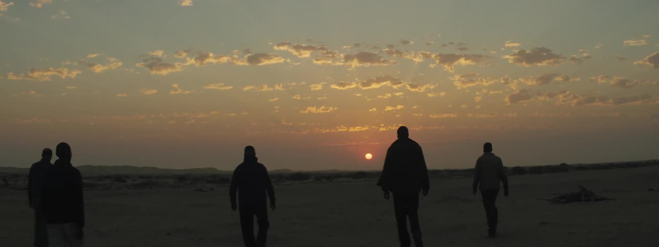 Sun sets in a cloud scattered sky as several people walk towards the sunset