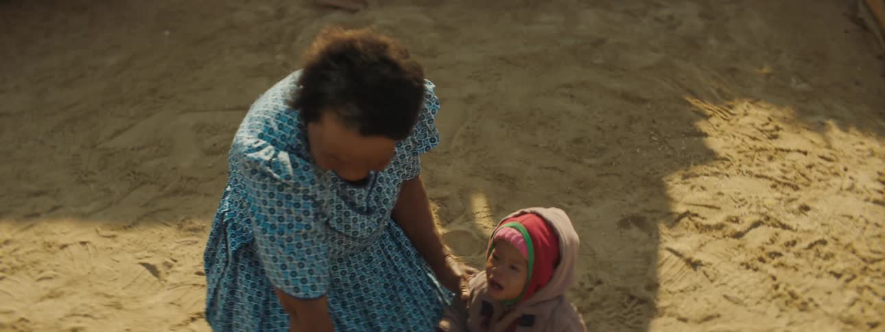 Man points a rifle at a woman kneeling on the ground next to a baby and a corpse