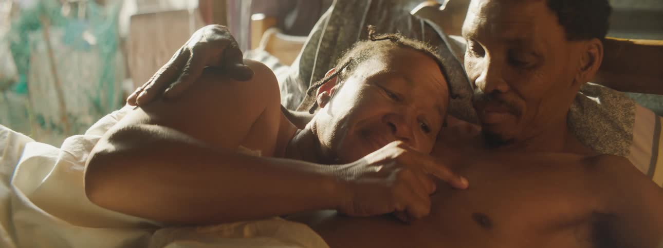 A black couple is in bed as the woman touches the mans bare chest