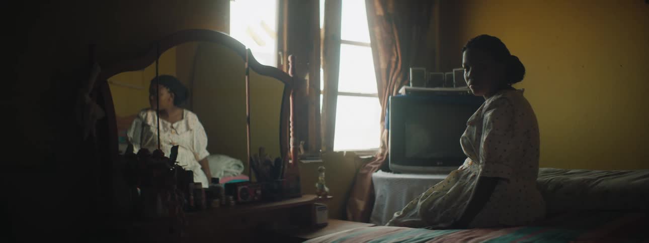 Woman sitting at the foot of her bed staring emotionlessly into mirror