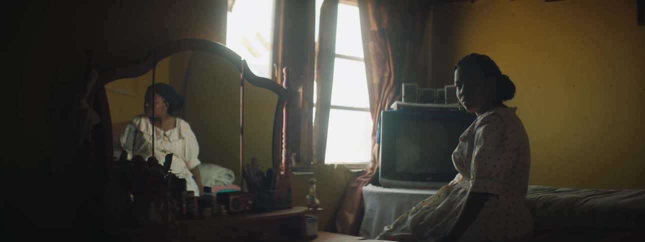 A young black woman sits on her bed with her head down and then looks to the side