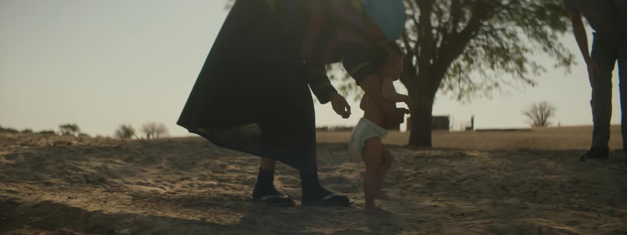 A black woman helping her baby girl learn how to walk outside with fathers encouragement in the distance