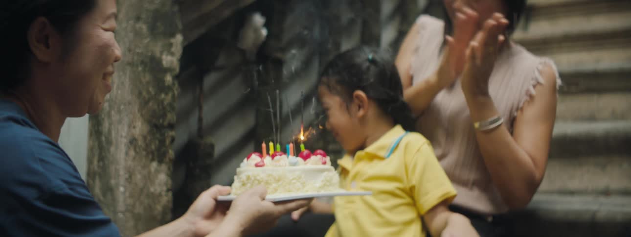A girl sitting in her mothers lap blows out the candles on a cake being held in front of her by her grandmother