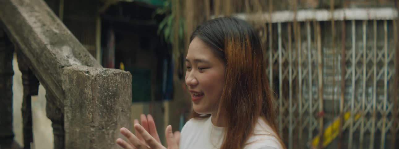 A young asian woman claps and sings while standing near a cement staircase outdoors during the day