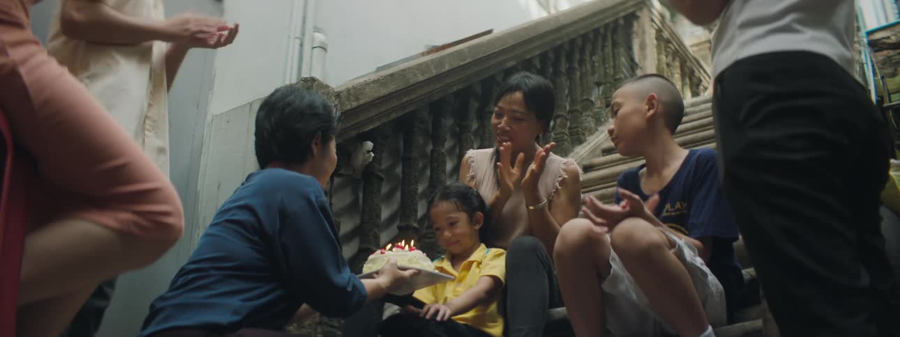 A man holds a birthday cake with lit candles in front of a little girl as she blows them out while surrounded by her clapping family