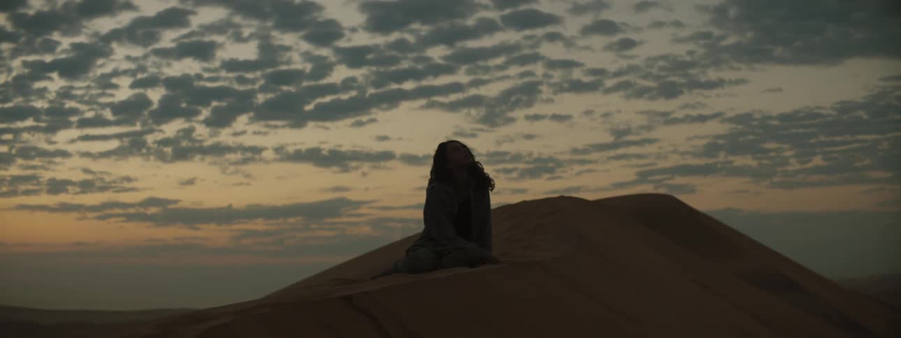 Woman sitting on sand dune is silhouetted against the sky in the desert at dusk