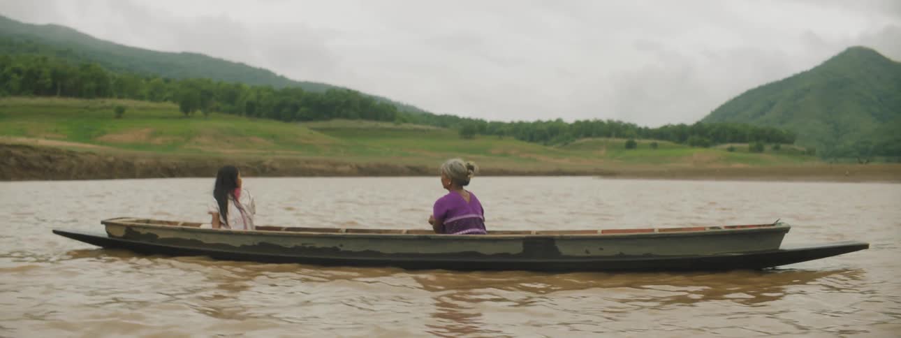 Two women sit in a boat on a lake as one of them uses an oar to move
