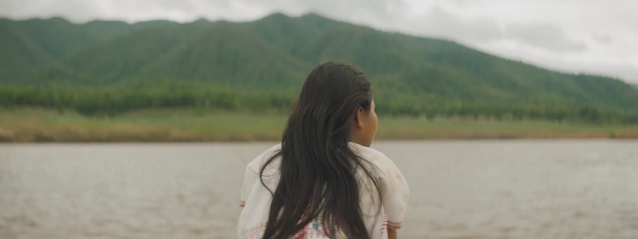 A girl gazes at the countryside around her as she rides down a murky river at the head of a boat