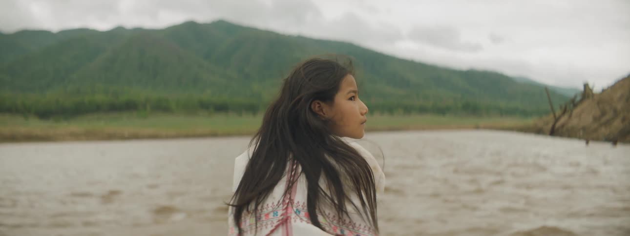 A girl looks around at her surroundings while riding at the front of a boat traveling down a murky river on a cloudy day