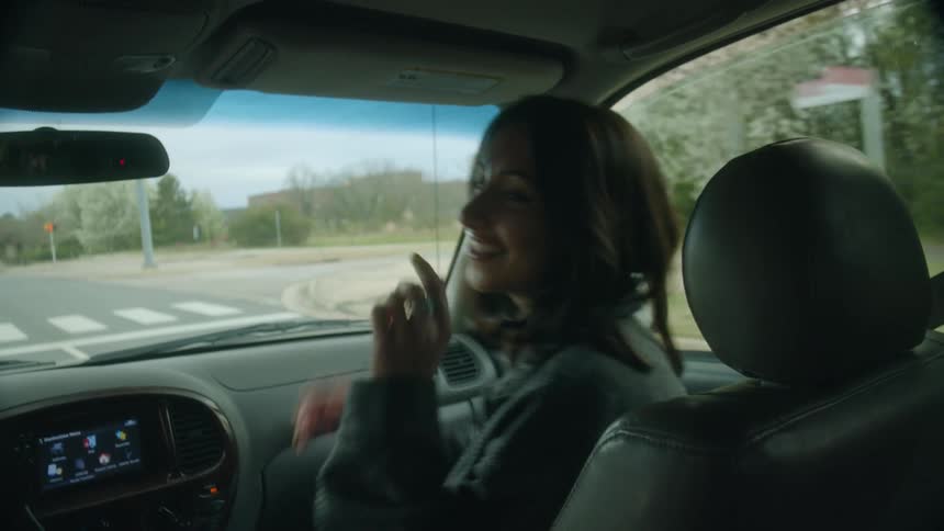 A woman laughs and dances in the passenger seat of a vehicle as it travels down a road during the day