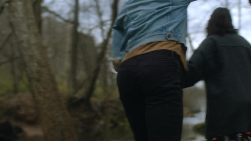 A teenage boy and girl hold hands as they walk towards a river in a forest
