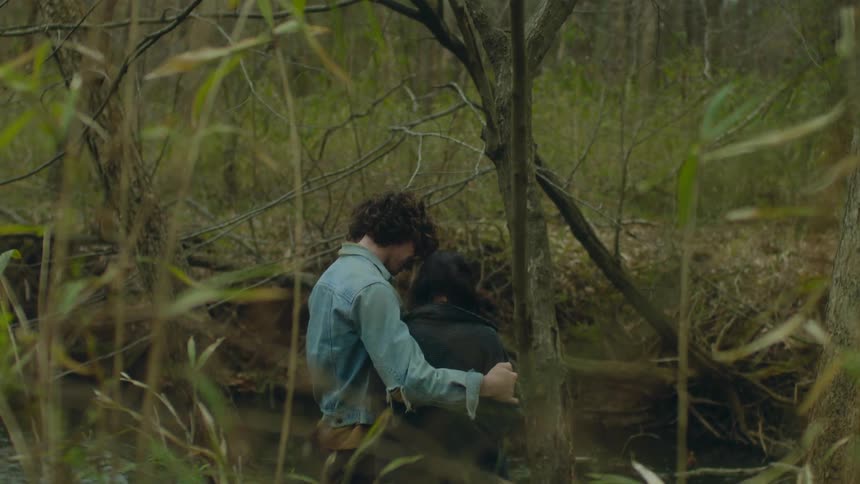 A teenage boy stands by a river with his arm around a girl and he rubs her back