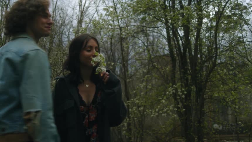 A man picks a twig of flowers from a tree and gives it to a young woman who gags upon sniffing it