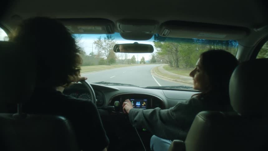 A teenage boy drives a car and a girl turns up the radio and they both move their head to the music and she laughs