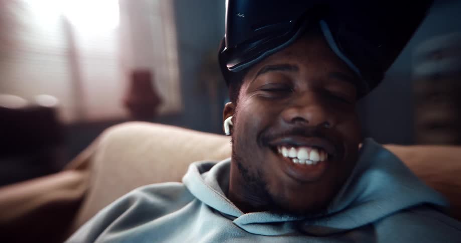 A man is sitting on a couch smiling and talking and he pulls his virtual reality goggles down over his eyes