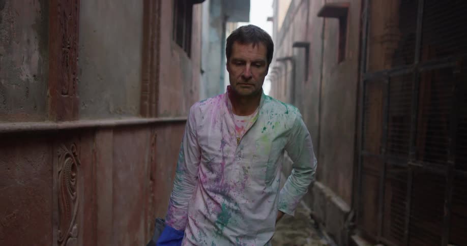 A man walks through a narrow alley and he's wearing a shirt stained with many colors