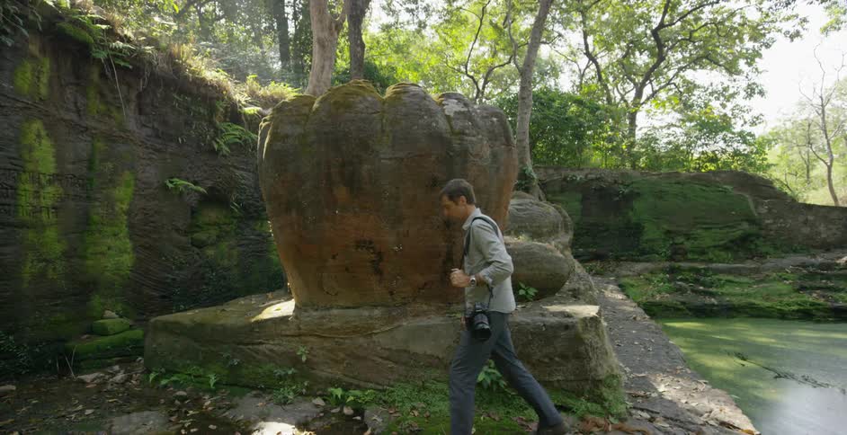 A man with a camera walks near some artifacts in a natural setting