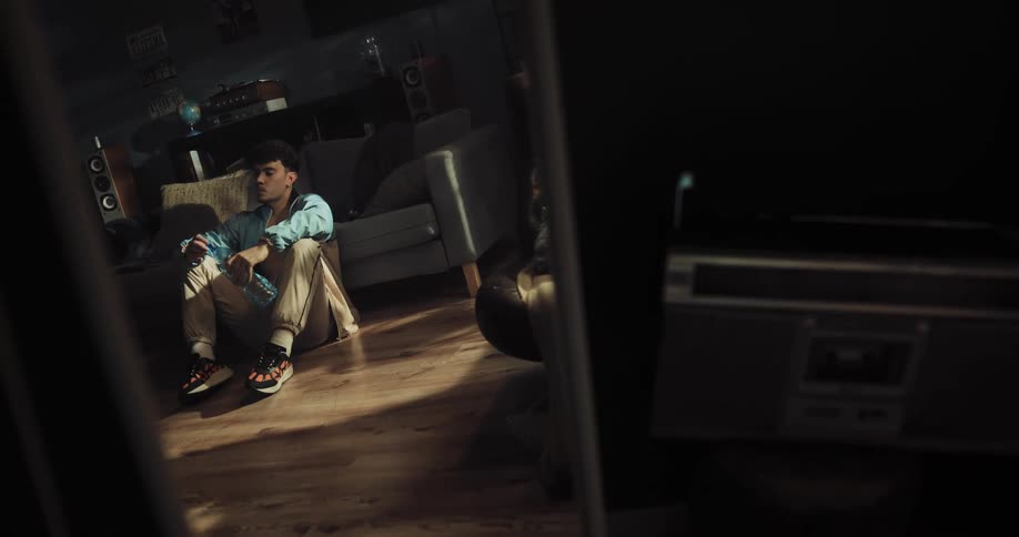 A man drinks from a water bottle before closing his eyes and sighing while sitting on the floor of a home living room during the day