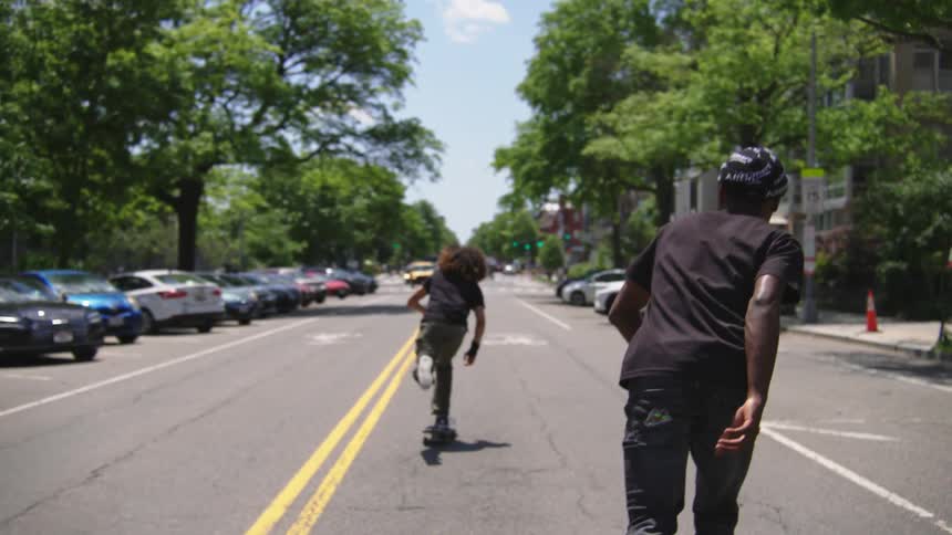 Two teenagers are riding skateboards down a street in light traffic and there are cars parked on the sides of the road in front of buildings and trees