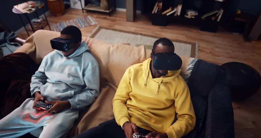 A pair of men play virtual reality games while sitting next to one another on a sofa in the living room of a house