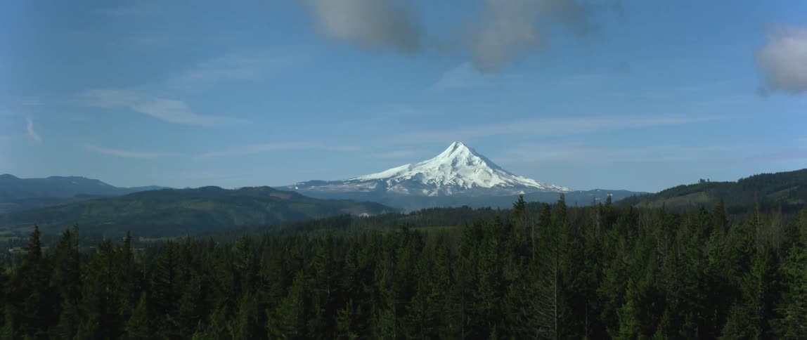 Farmland is amongst the forest in a mountain range with the tallest mountain having a snow cover