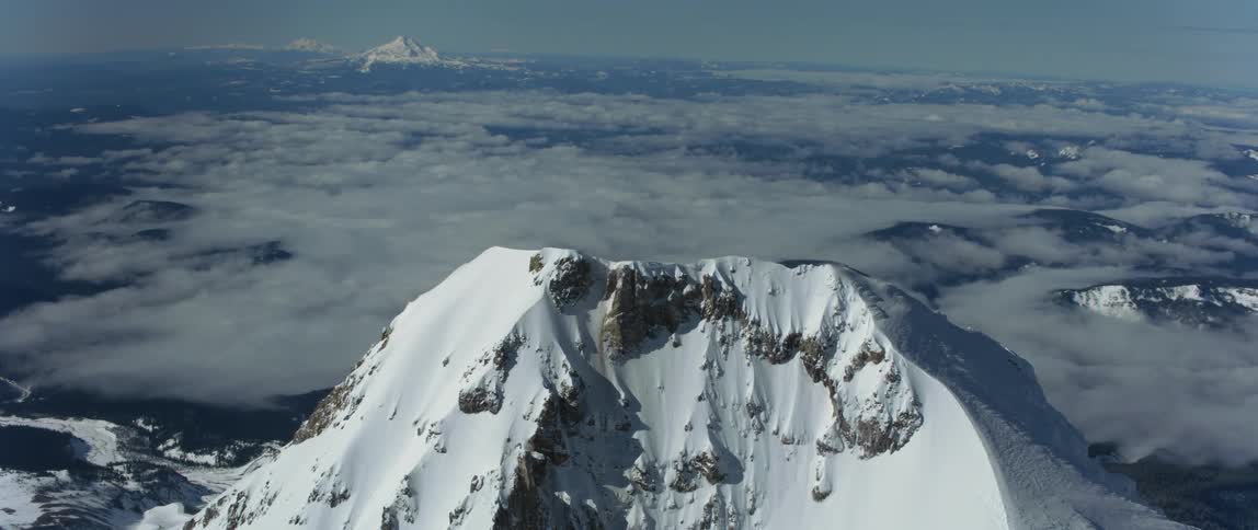 A snow capped mountain peak looms over a vast countryside obscured by clouds