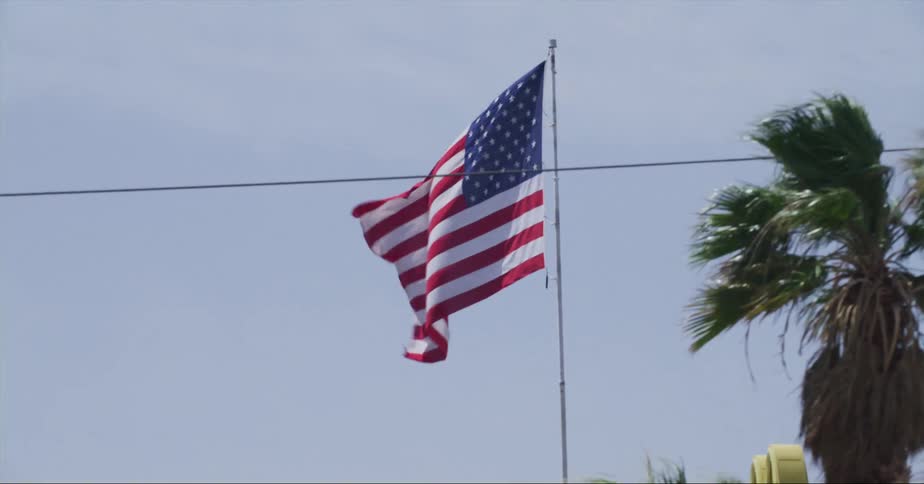 An american flag on a pole waves in the sky above a street lined with palm trees