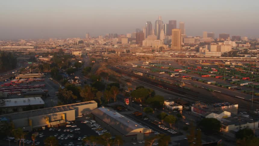 An aerial view of cars and shipping trucks driving a highway past a lot filled with shipping containers with a city skyline with offices in the distance