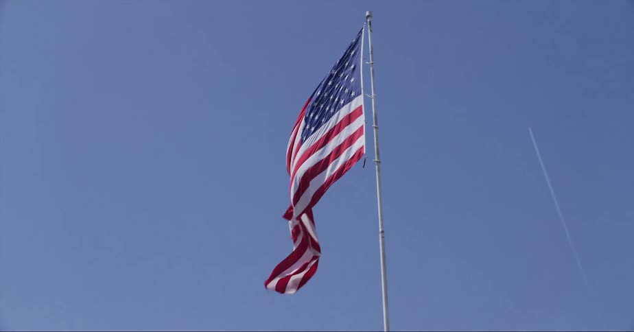 American flag flutters in the wind near street corner with contrail in the background