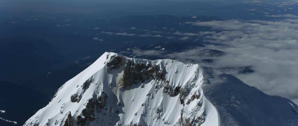 The sun shines on a snow covered mountain ridge high above the clouds and there are snow covered mountains in the distance