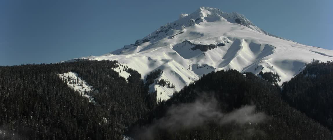 Fog rises above a forested mountain with a snow covered peak high in the clear blue sky