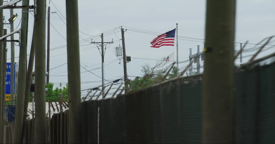 An american flag on a pole flying and waving above a fence near power lines