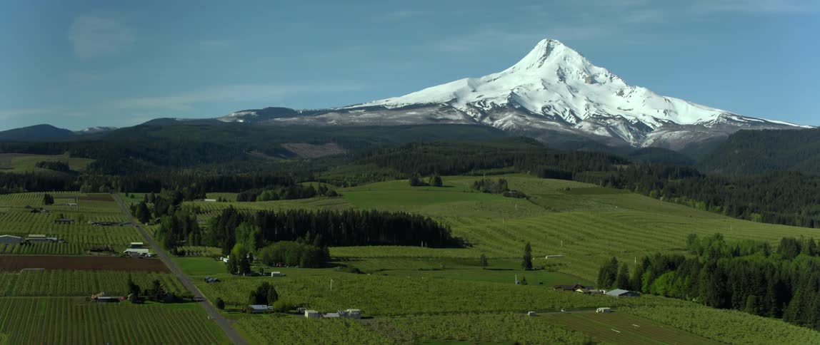 A snow capped mountain peak looms over verdant farmland in the countryside during the day