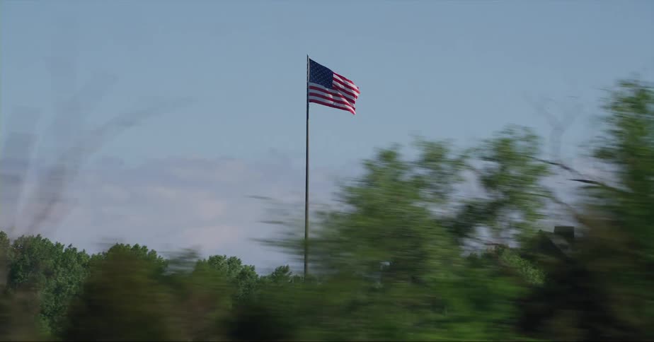 An american flag waves in the breeze on a pole above trees and next to a building