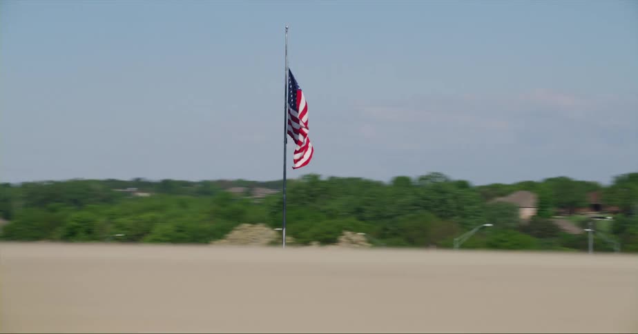 An american flag waves in the wind on a pole on the roadside as traffic passes by and there are buildings and trees in the distance