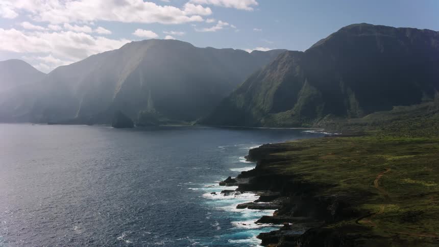 Waves roll in crashing against rocks along the shoreline and there are mountains in the distance under a cloudy sky