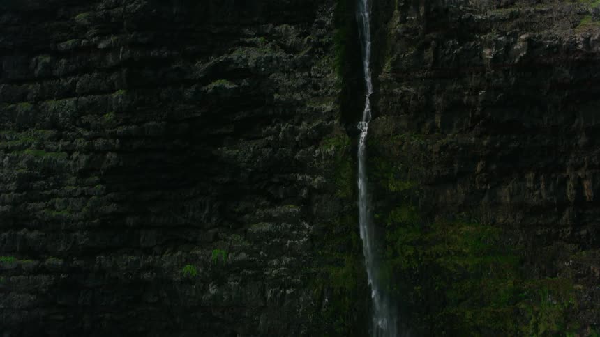 Long thin waterfall flows down seaside cliff onto ground as waves crash against narrow shore