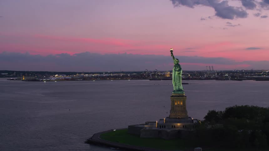 The statue of liberty overlooks new york harbor as boats sail across the water under a bright pink sky