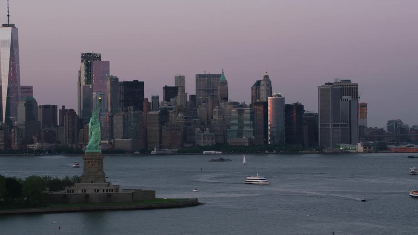 The statue of liberty overlooks new york harbor as boats sail across the water and tall city buildings are in the distance