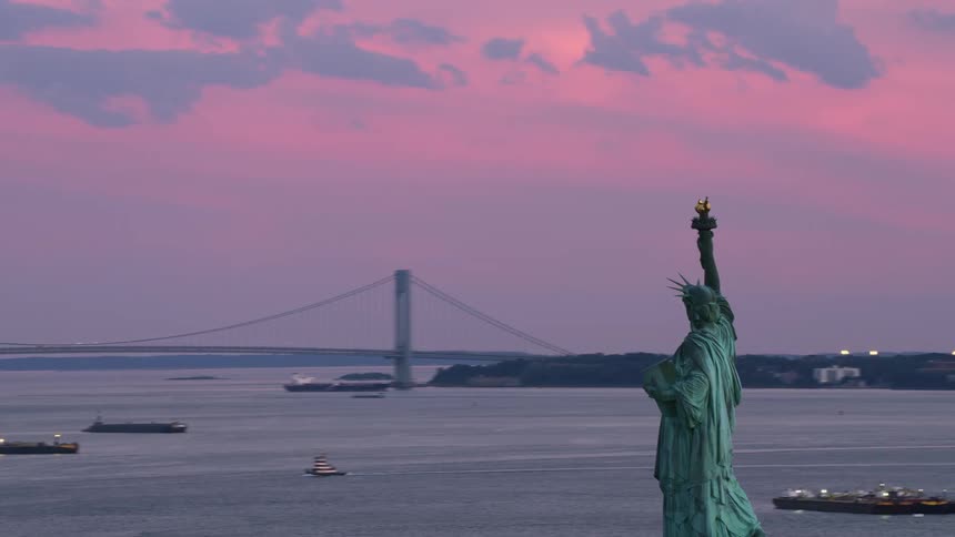 The statue of liberty overlooks new york harbor as boats sail across the water under a pink sky