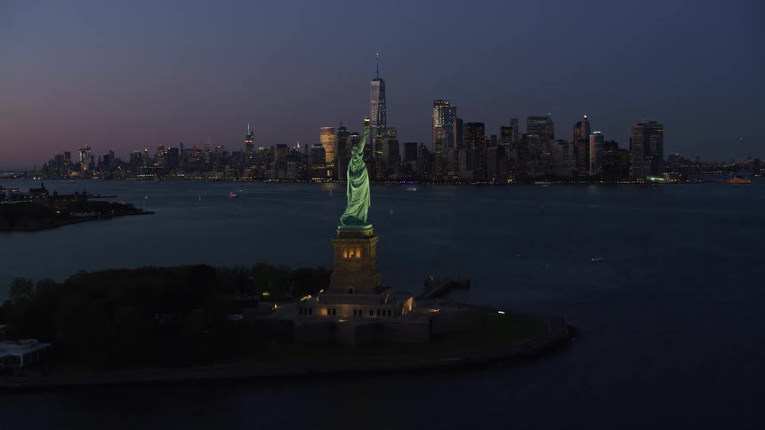 The statue of liberty overlooks new york harbor as boats sail and lights illuminate city buildings in the distance