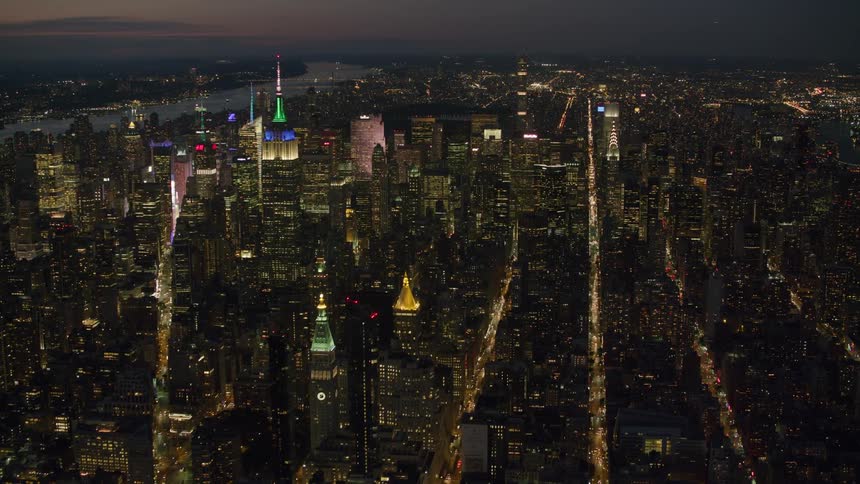 The spire of the empire state building is colorfully lit above new york city at night