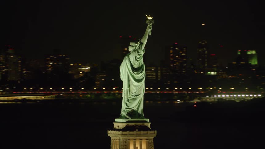The statue of liberty overlooks new york harbor as lights illuminate city buildings in the distance