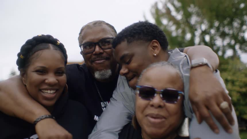 Family hug each other and smile at barbecue with grill in the background