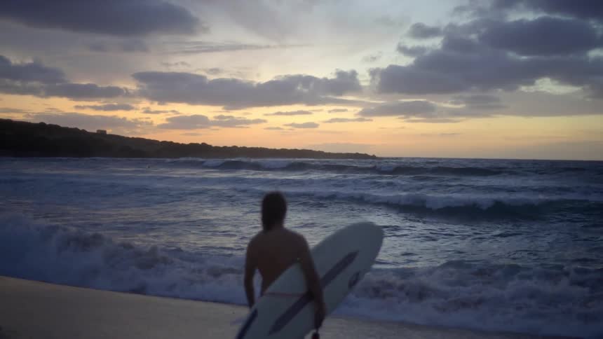 A male stands on the beach while holding a surfboard while watching the ocean waves