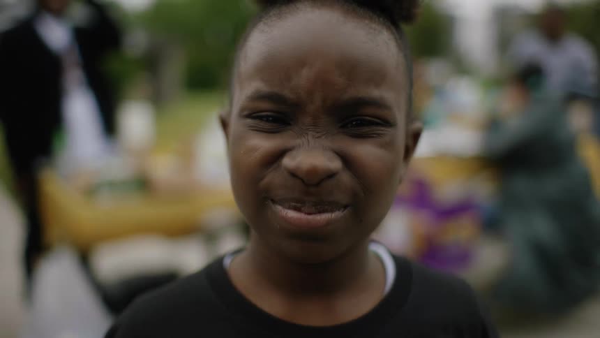 A girl closes her eyes and makes a funny face while family mills around behind her