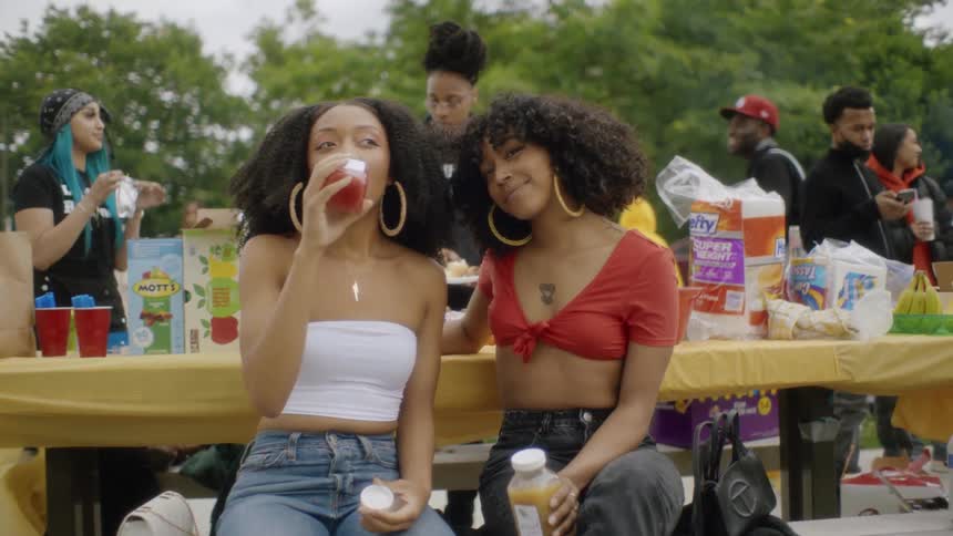 Two teenage girls sit on a picnic bench in a park drinking juice and hug and smile as other behind them wander around drinking