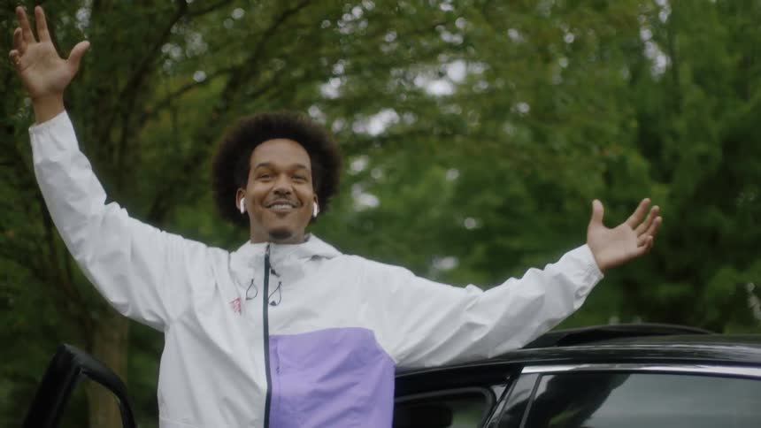 A man gets out of his car smiling with arms raised greeting people