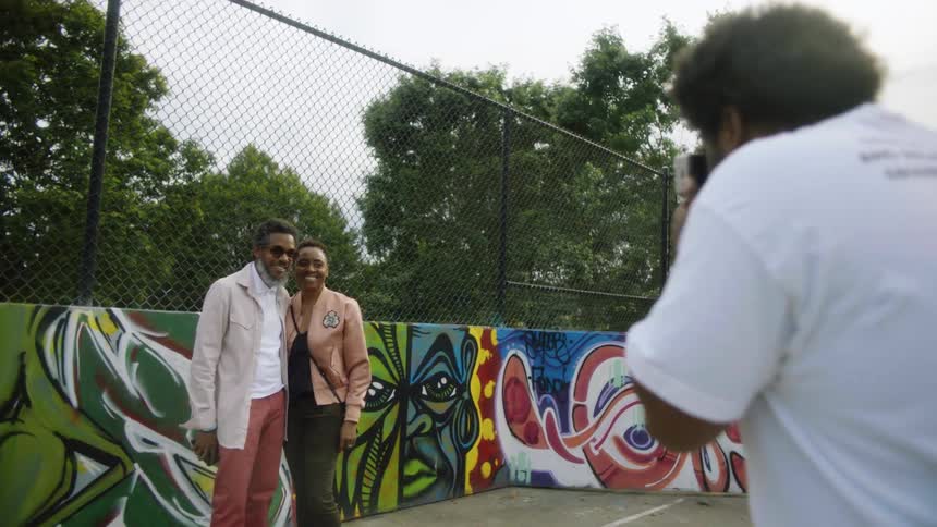 A photographer is taking a picture of a man and a woman standing in front of artwork near a fence at a park