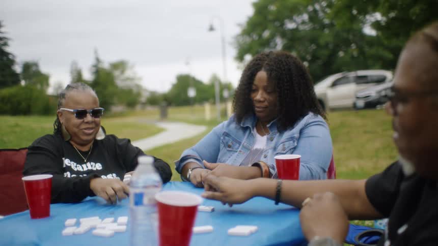 Family sits around table in park playing dominoes